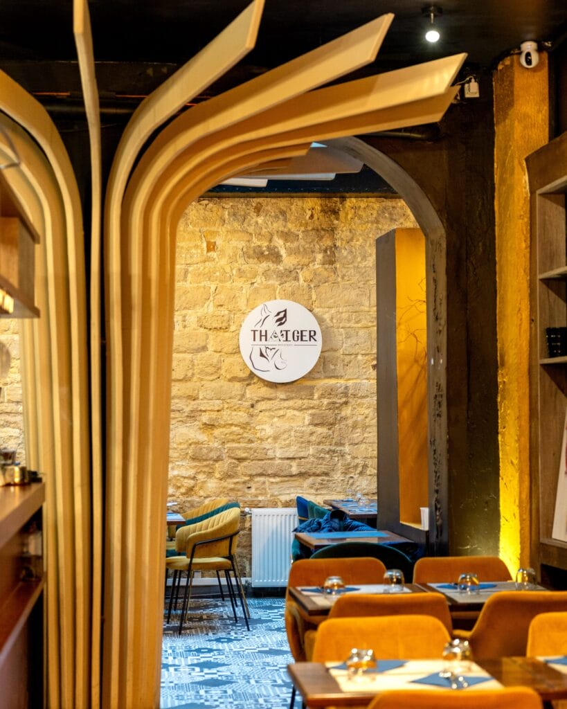 Un intérieur de restaurant moderne avec des arches en bois, des murs en pierre beige, des chaises élégantes, des tables dressées et une enseigne murale circulaire « THATGER » avec des illustrations minimalistes. Un éclairage d'ambiance chaleureux crée une atmosphère chaleureuse.